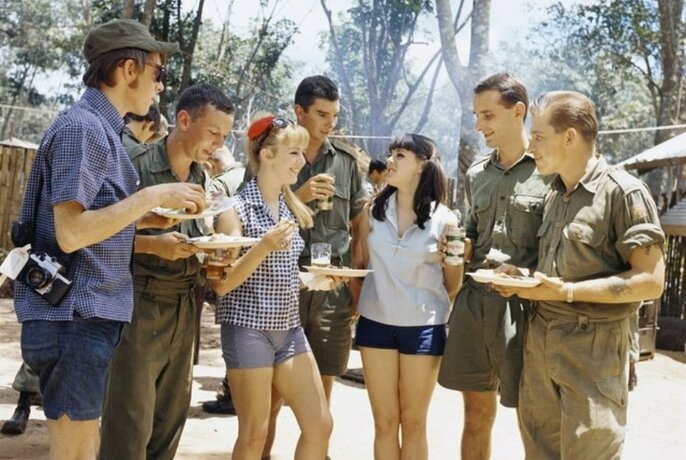 Two women in shorts at centre of small group of soldiers, all holding plates of food and drinks.