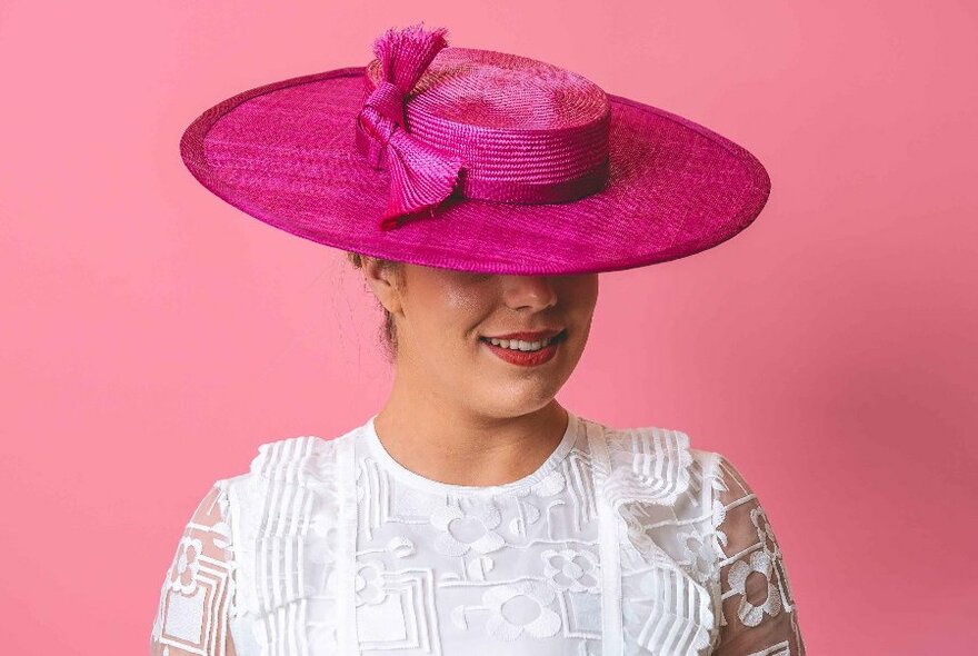 A model wearing a frilly white shirt and a bright pink boater-style hat, against a pink background.
