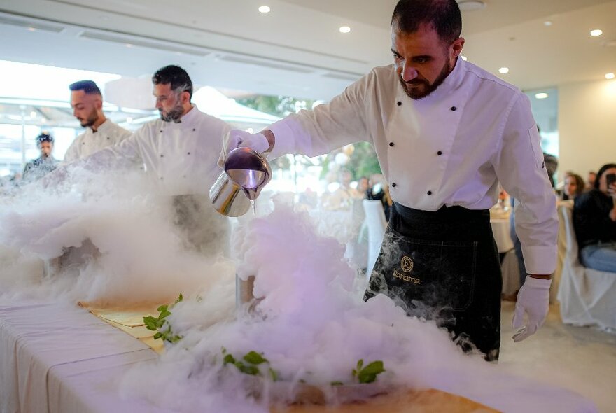 Chefs in white uniforms pouring water to create steam onto dishes of food.