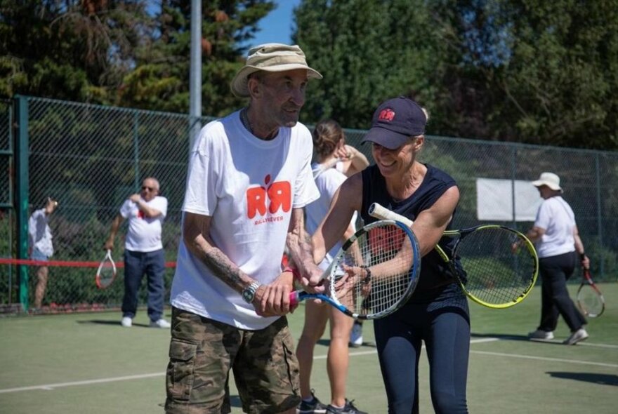 Someone being shown how to hold a tennis racquet by an instructor outdoors on a tennis court with others in the background. 