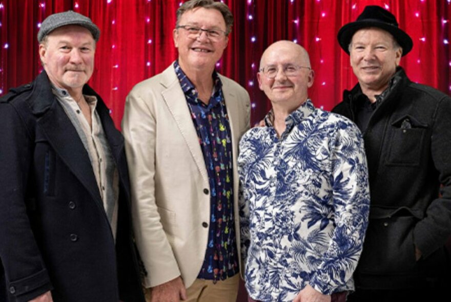 Four older male musicians post in front of a red curtain with fairy lights. 