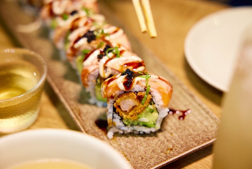 A close up of sushi rolls featuring crispy prawns, avocado and kewpie mayo - a set of chopsticks about to dive in.