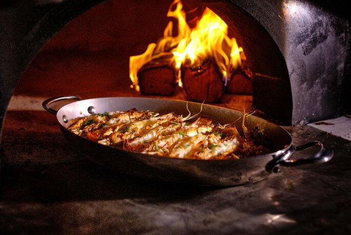 Metal platter of cooked prawns resting on a table in front of an open wood-fired oven.