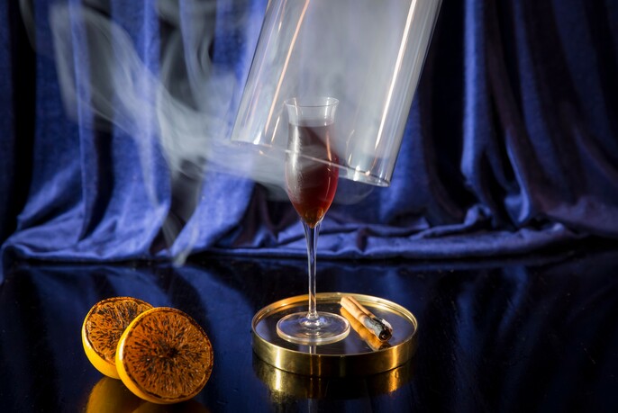 Small glass of liquor under a bell jar containing burnt orange and cinnamon smoke.