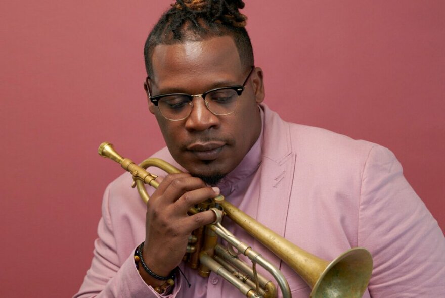 Trumpet player Keyon Harrold, holding his instrument and wearing a pink suit, against a darker pink background.