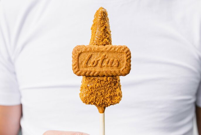 A biscuit covered wedge of cheesecake on a stick held by someone wearing a white tshirt. 