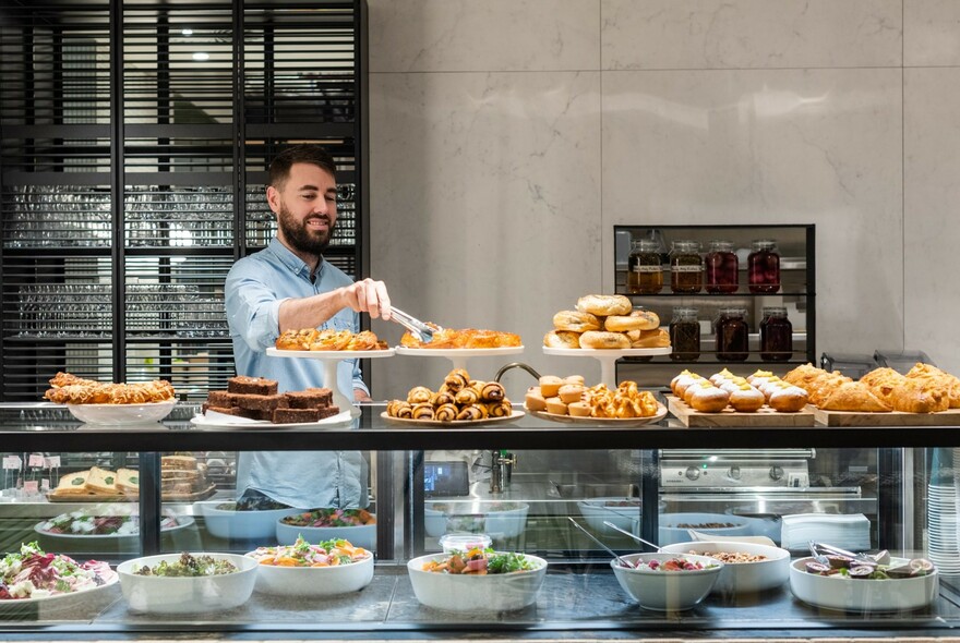 Man arranging pastries on a cafe display counter that also contains bowls of salad.