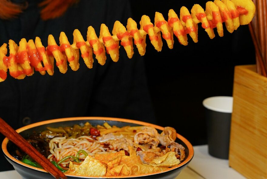 A bowl noodles and vegetables, chopsticks resting on the side and a twisted long noodle being held up above the bowl.