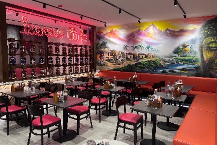 Brightly decorated restaurant interior, with a large mural on the rear wall, small square black tables and red cushioned chairs.