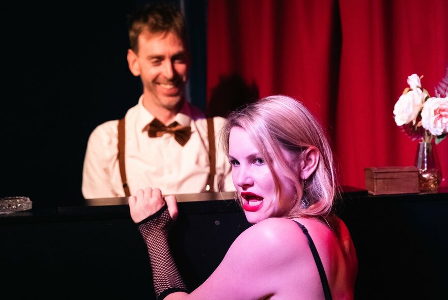 A woman performs in a cabaret setting with a pianist accompanying her and laughing.