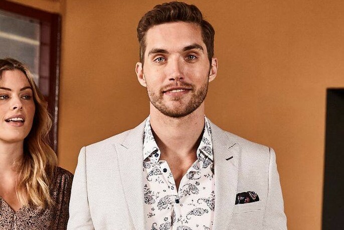 Smiling bearded model wearing a linen suit jacket and paisley shirt, next to a woman in a doorway.