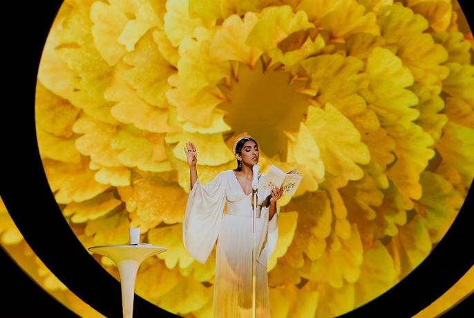 Poet Rupi Kaur wearing a long white dress, reading from a book in her hands, in front of a giant backdrop of a yellow flower.