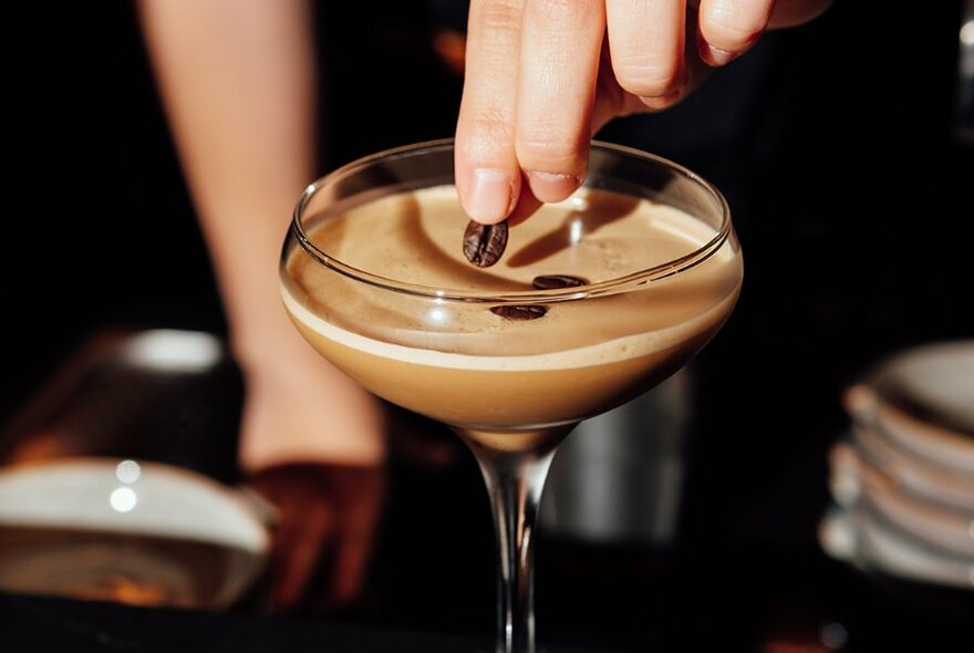 A hand placing coffee beans onto the surface of a cocktail.