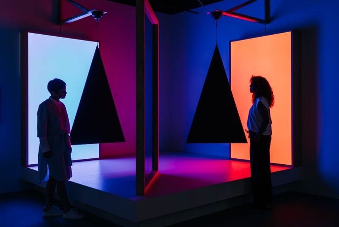 Two people standing in a darkened room with large dark triangles hanging in front of them, coloured screens behind.