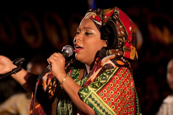 Woman wearing a colorful patterned dress and headscarf singing into a microphone. 