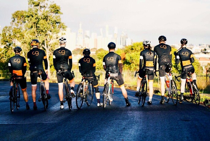 Rear view of eight cyclists in lycra riding gear straddling their bikes on a wide bend in the road.