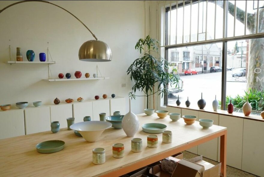 A wooden table in a shopfront with ceramic pots and plates arranged around the walls, window ledge and table.