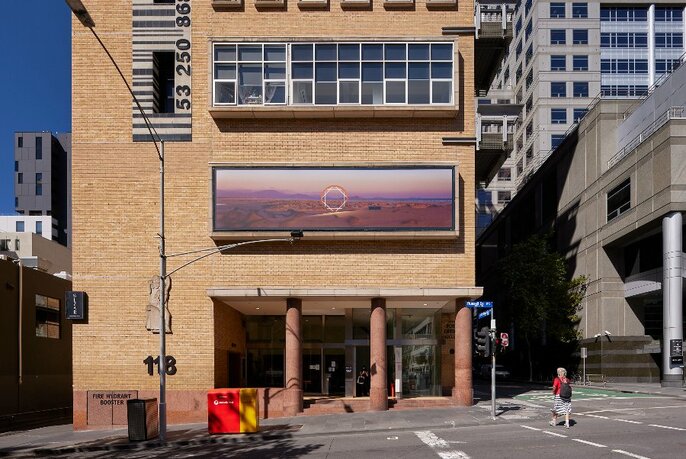 Photographic artwork installed on the exterior of a building, with other buildings, pedestrian and a road also visible.