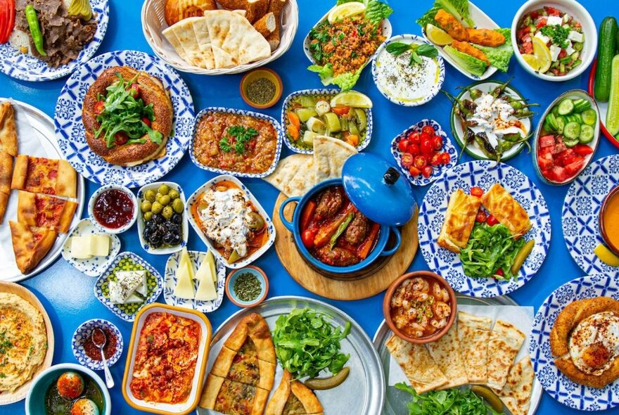 Overhead view of many Turkish banquet dishes on a blue tablecloth including salads, dips and breads.