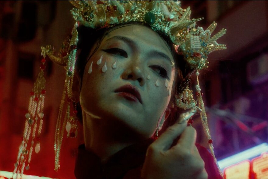 An Asian woman in elaborate golden headdress and wax tear drops on her cheek.