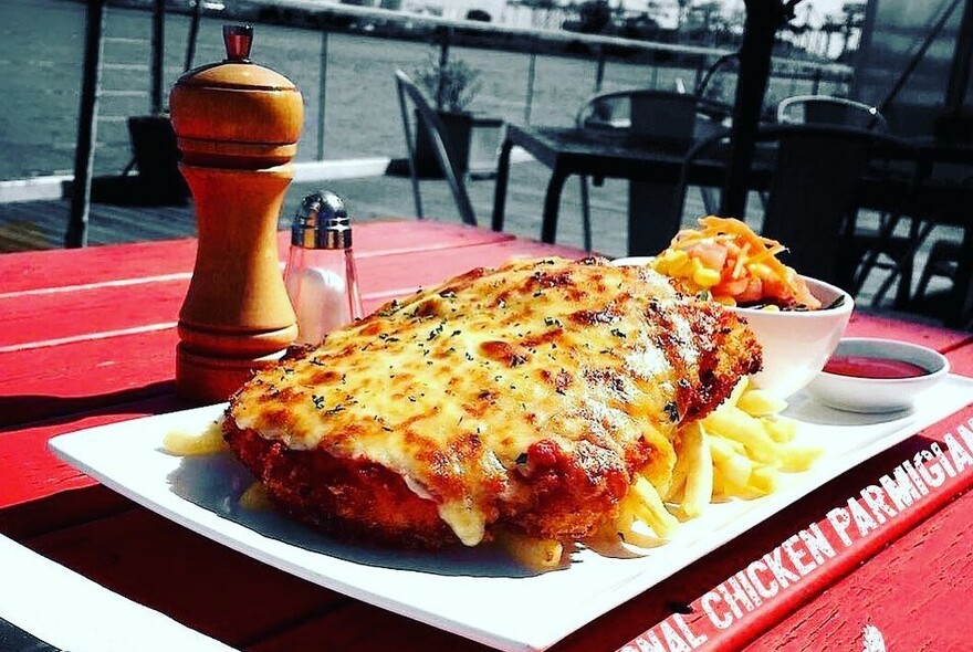Outdoor table set with a red tablecloth, huge chicken parmigiana on a white platter, chips and condiments.