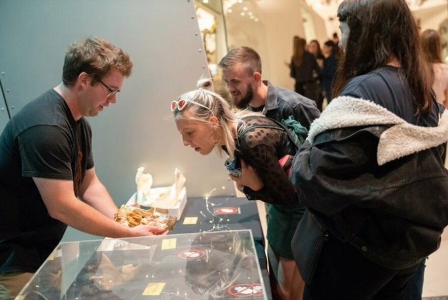People looking at objects a man is holding on top of a glass display case in a museum.