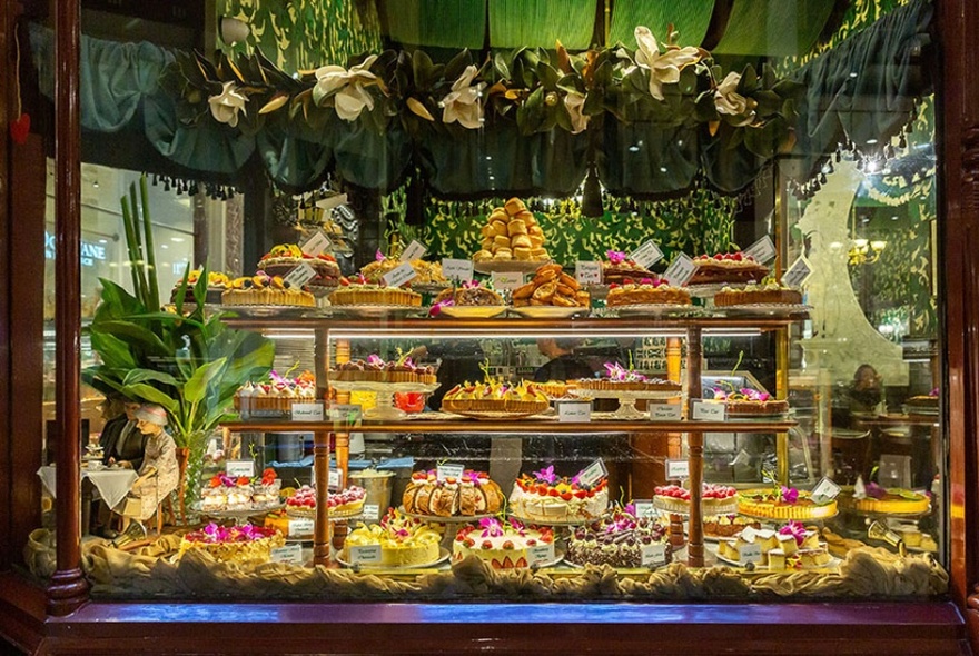 Large glass exterior window of a cafe showing three levels of assorted cakes and desserts on display, surrounded by floral arrangements.