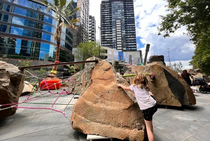 Child clambering over large rocks in a city playground space with skyscrapers visible in the background.
