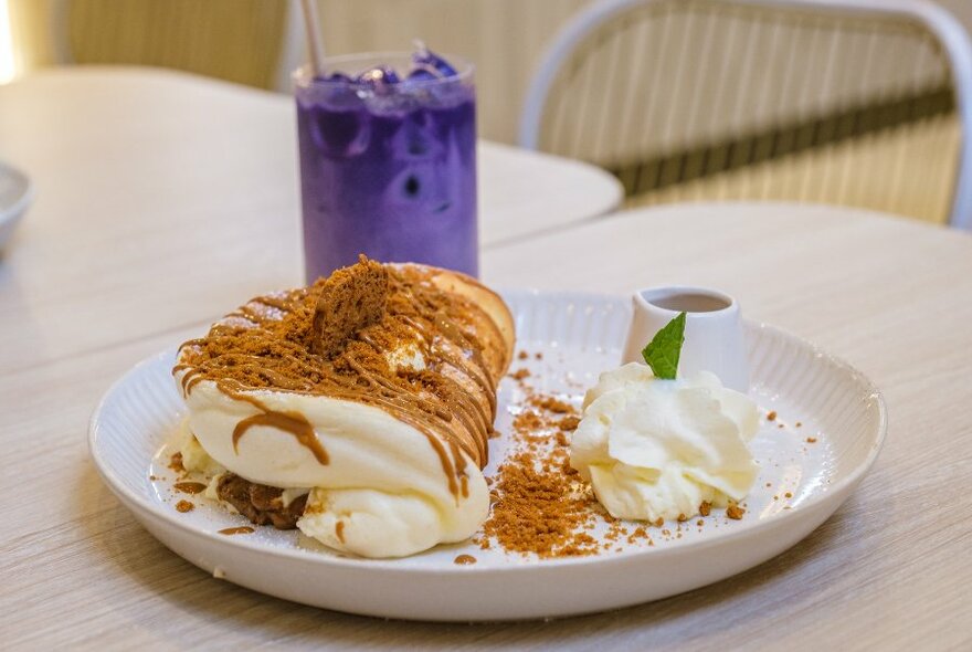A plate with a soufflé pancake folded over with chocolate biscuit crumbs and whipped cream with a tall purple drink. 