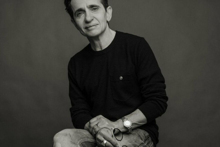 Journalist Masha Gessen looking thoughtful, wearing a black top, sitting with one leg crossed over the other, holding reading glasses in their hands; black and white image.
