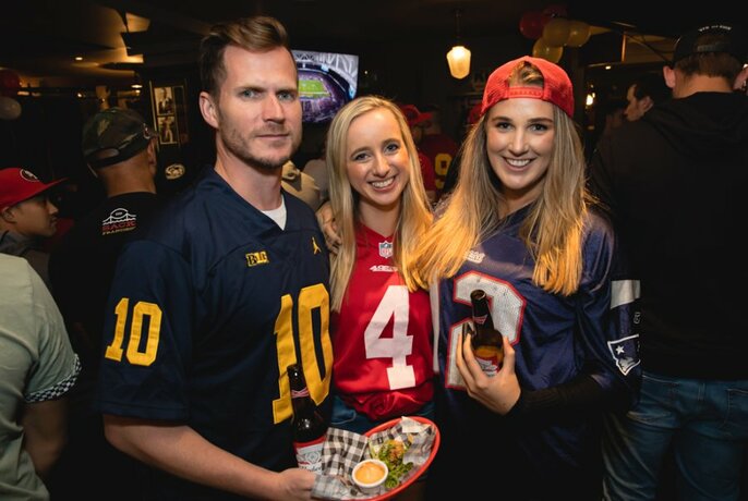 Three smiling people in a pub interior wearing sporting club jumpers, one holding a beer and another one a plate of food.