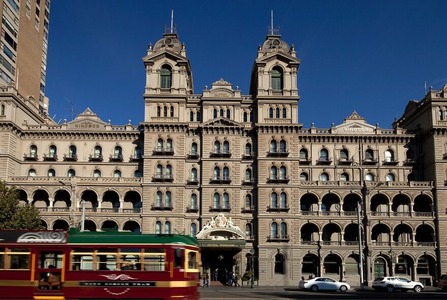 Grand facade of the 140-year old Windsor Hotel against a blue sky, with a tram shuttling past on the road.