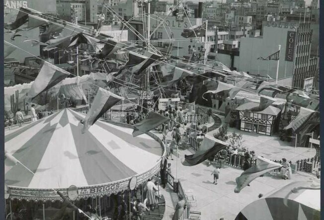 Black and white photo of a carnival held on a rooftop featuring a Ferris wheel, carousel and bunting flags 