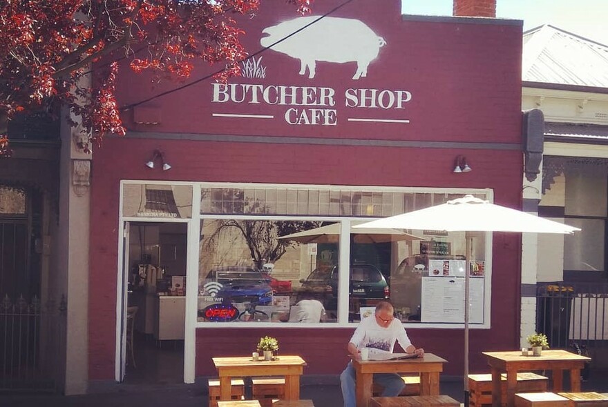 Person sitting at small table under umbrella outside Butcher Shop Cafe.