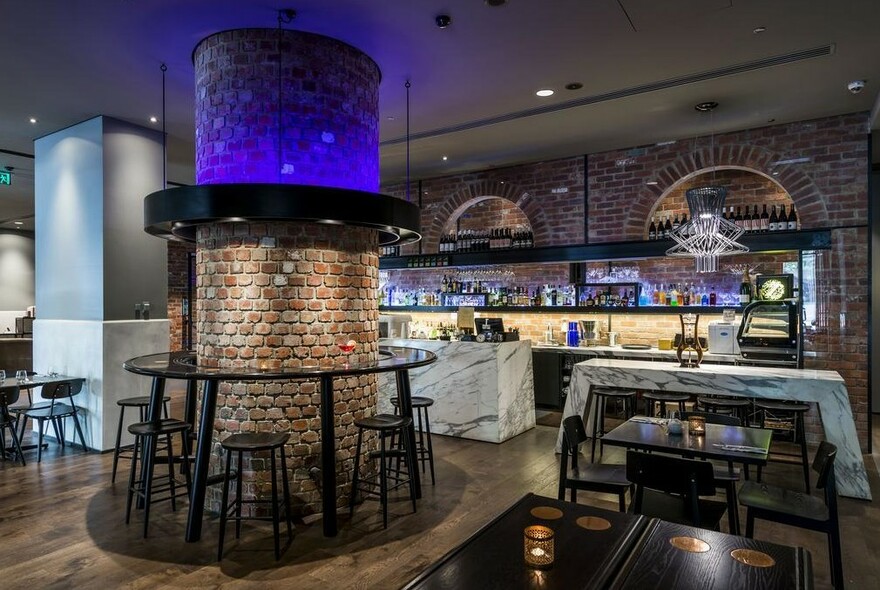 Moodily lit restaurant interior with central brick pillar surrounded by stools, backlit cocktail bar in the background.