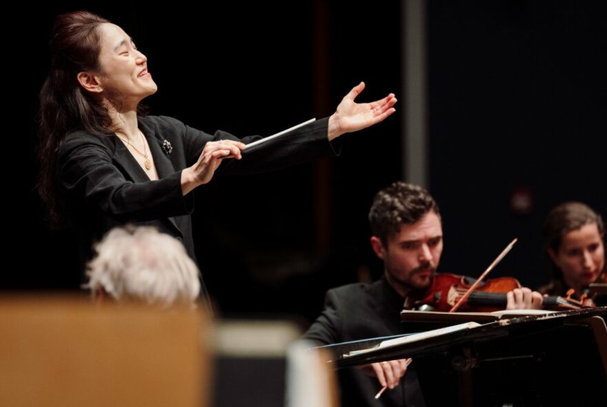 A conductor beams as she holds her hand out with two violinists visible. 