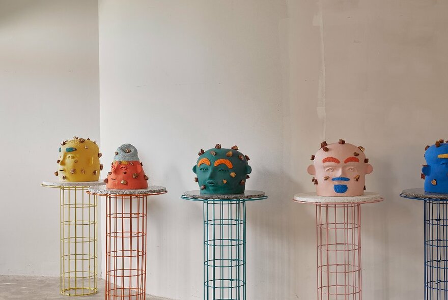 Small bright sculptures of fantasy heads on display on raised plinths, against a white wall.