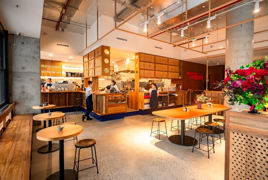 Spacious cafe interior with a central shared wooden table with stools surrounded by wooden cabinets, concrete floor and a kitchen service area at the back.