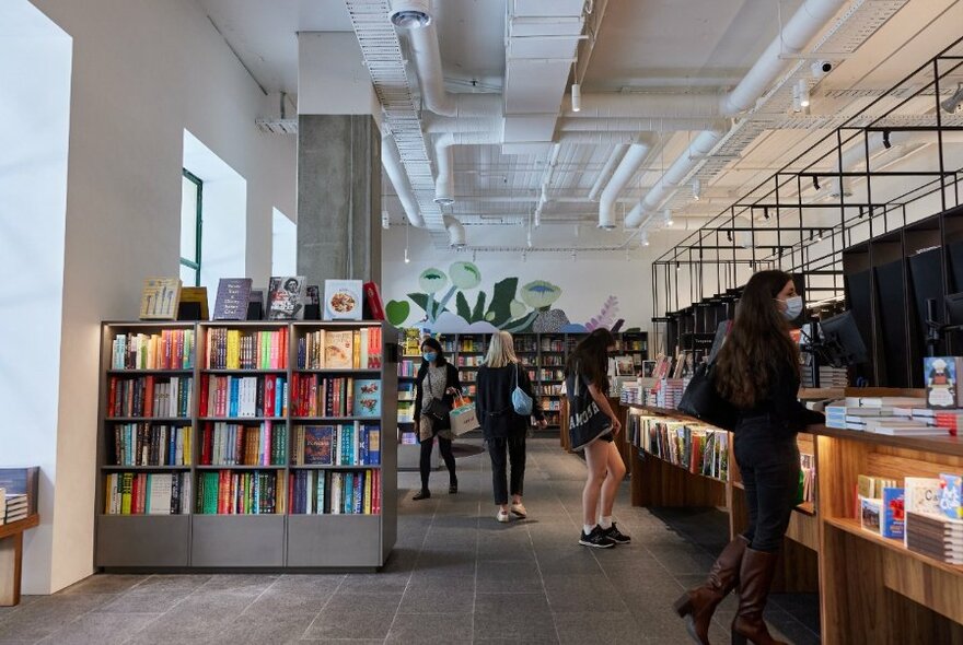 Interior of bookshop with shoppers walking around, display shelves of books and plant greenery in the background.