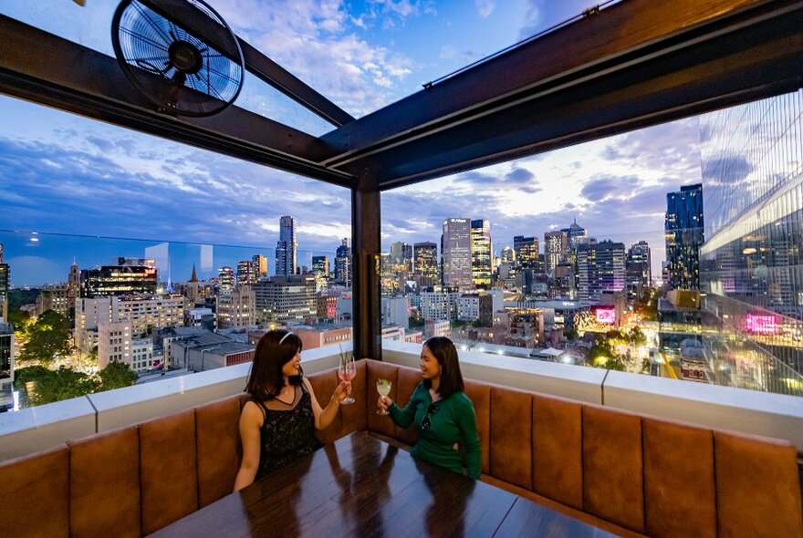 Two girls drinking cocktails in the corner booth of a rooftop bar overlooking the city skyline.