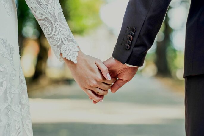 A close up of the clasped hands of two people wearing bridal wear. 