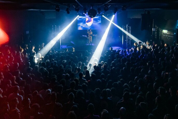 White search-like spotlights being projected onto a crowded dance floor with a performer on the stage in the background standing at a DJ mixing desk. 