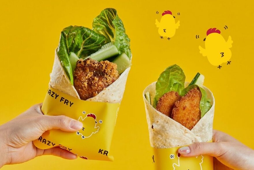 Two hands holding crispy fried chicken wraps with lettuce and cucumber, wrapped in bright yellow paper packaging, against a yellow background.