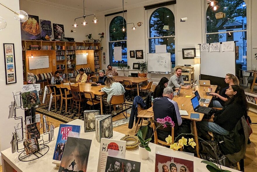 A large airy studio space with adults seated around several tables writing on their laptops or in journals, the walls surrounded by shelves with books on them. 