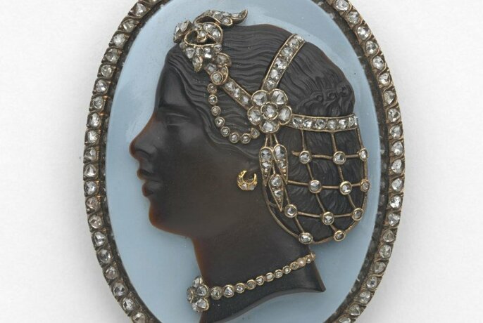 Cameo brooch depicting the profile of a head wearing a jewelled hairpiece, necklace and jewels in the border edging of the cameo.
