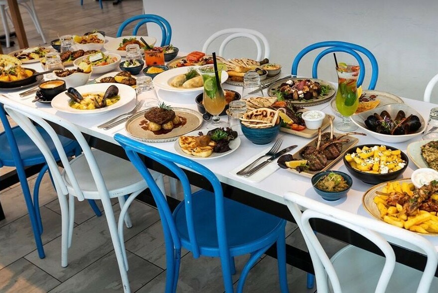 A Greek table with white and blue chairs set with an array of dishes, bowls and cocktails.
