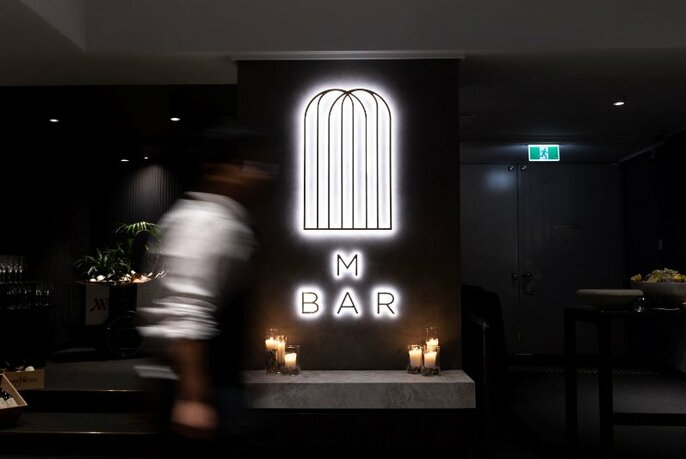 A blurred waiter walking past M Bar signage with candles.