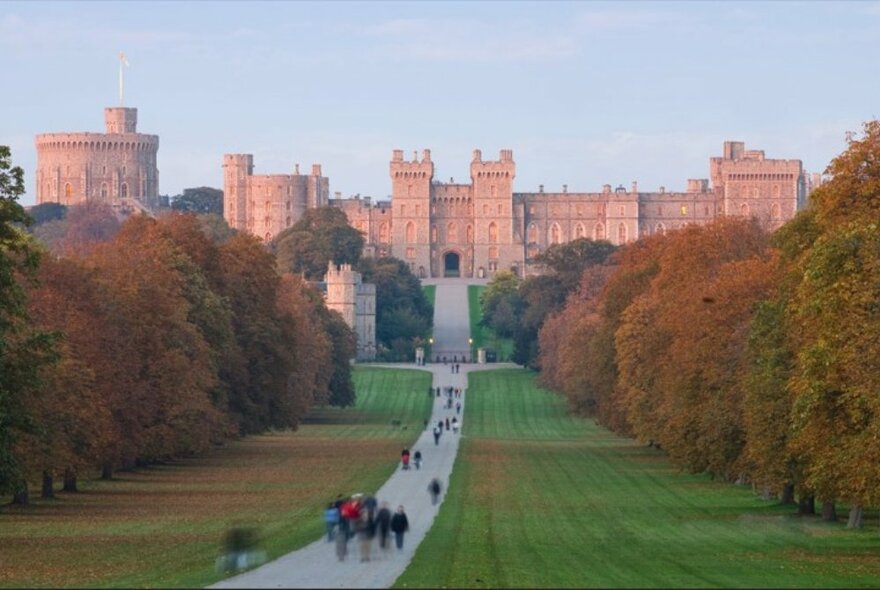 Windsor Castle at sunset, with people walking up the long path through the green fields in front of the castle.