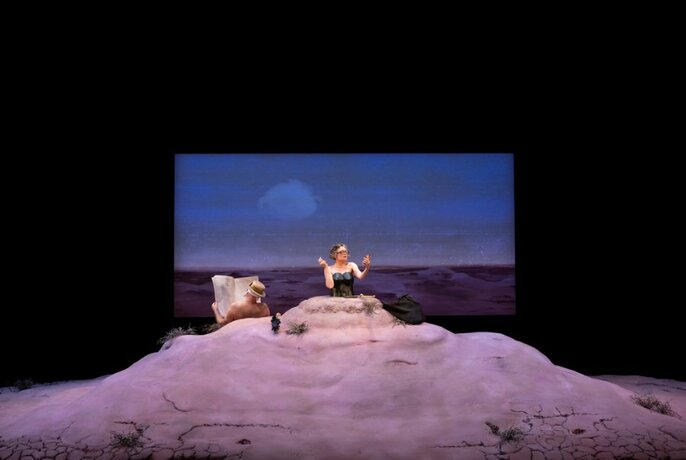 Performer Judith Lucy from the waist up, the lower half of her body buried in a small hill, on a stage with a landscape background displayed on a screen behind her.