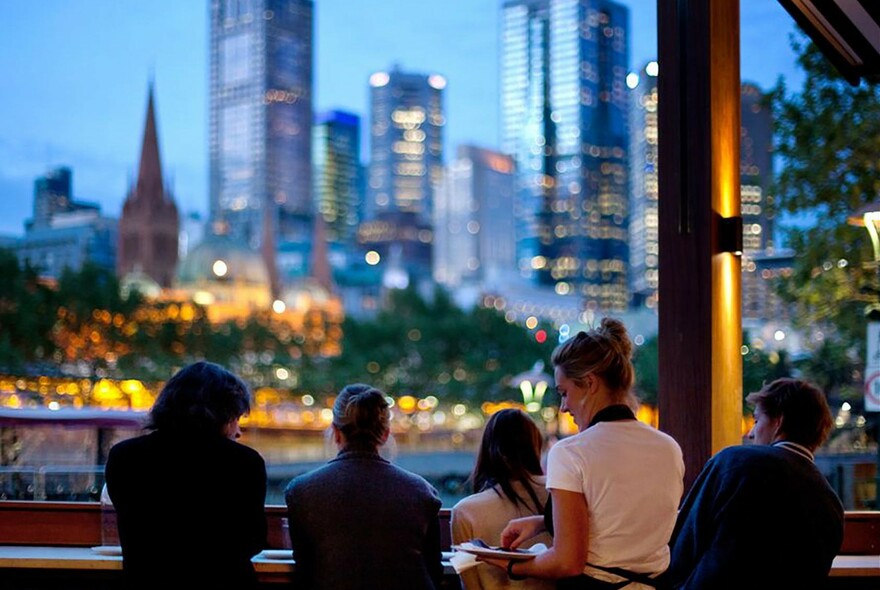 People seated at a bar with a waitress, looking over to twilight-lit city buildings in the distance.